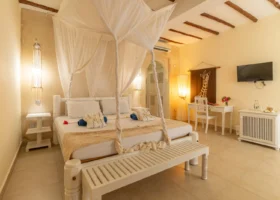 Interno di una camera da letto in stile afro-chic al 7 Islands Resort di Watamu. Al centro spicca un grande letto matrimoniale in legno bianco con zanzariera a baldacchino in tulle leggero, asciugamani azzurri e fiori di frangipane decorativi. Sullo sfondo pareti color sabbia, soffitto con travi in legno a vista, una scrivania bianca con sedia intagliata, un quadro con giraffa e un televisore a schermo piatto.