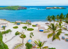 Panorama dall'alto della spiaggia corallina del 7 Islands Resort a Watamu. La sabbia bianca è punteggiata da piccoli arbusti verdi, palme, divani in legno con cuscini celesti e gazebo prendisole. L'oceano Indiano sfoggia intense sfumature di blu e turchese, interrotto all'orizzonte dai caratteristici isolotti verdi che danno il nome al resort.