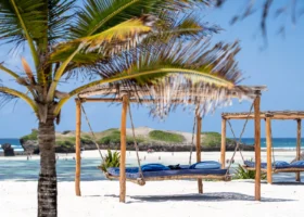 Primo piano di un letto oscillante sospeso in legno con materasso blu e cuscini azzurri, posizionato sulla sabbia bianca della spiaggia del 7 Islands Resort a Watamu. Una palma parzialmente sfocata incornicia la scena a sinistra. Sullo sfondo si scorgono la barriera corallina, l'oceano indiano turchese e un atollo verdeggiante durante la bassa marea.