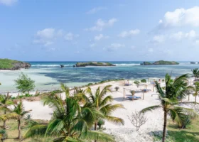 Vista panoramica sulla costa di Watamu fronteggiante il 7 Islands Resort. La spiaggia bianca degrada dolcemente verso fondali bassi e trasparenti, tipici del fenomeno della bassa marea, svelando venature di alghe e sabbia. Strutture balneari in legno e ombrelloni si integrano nel paesaggio tropicale costellato di palme e scogliere coralline sullo sfondo.