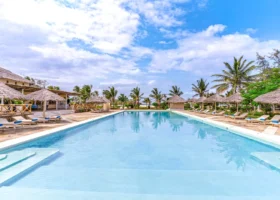 Ampia piscina rettangolare con acqua azzurra e limpida al centro del solarium del 7 Islands Resort. La pavimentazione esterna in pietra ospita lettini in legno e ombrelloni in paglia disposti simmetricamente. Sulla sinistra si nota la struttura del bar ristorante con tetto in makuti, mentre sullo sfondo svettano palme verdi sotto un cielo parzialmente nuvoloso.