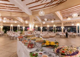 Interno del ristorante del 7 Islands Resort a Watamu durante il servizio a buffet. Un lungo tavolo centrale è allestito con insalate, piatti freddi, frutta tropicale tra cui ananas intagliati, e specialità di pesce. La sala presenta un'architettura aperta con imponenti colonne chiare, soffitto con travi in legno sinuose, lampadari sferici in corda intrecciata e tavoli apparecchiati con sedie bianche.