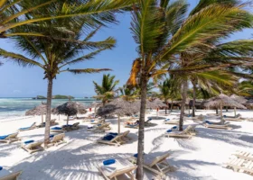 Spiaggia di sabbia bianca finissima del 7 Islands Resort a Watamu, Kenya, ombreggiata da alte palme da cocco. Numerosi lettini prendisole in legno con teli mare azzurri sono disposti sotto ombrelloni in paglia makuti. Sullo sfondo si estende il mare cristallino color turchese con atolli corallini affioranti sotto un cielo azzurro e limpido.