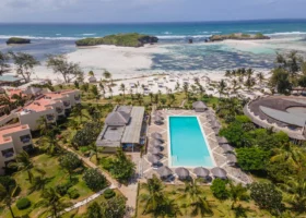 Prospettiva aerea perpendicolare che mostra il layout del 7 Islands Resort a Watamu. Al centro domina la grande piscina turchese circondata da ombrelloni circolari disposti a scacchiera. Attorno si sviluppano i giardini tropicali curati, i corpi di fabbrica degli alloggi con tetti piani sul lato sinistre, la reception circolare sulla destra e l'accesso diretto alla spiaggia e al mare cristallino sulla parte superiore.