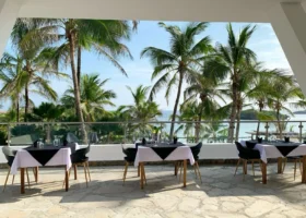 Interno dell'ampio ristorante del Barracuda Inn Resort in Kenya, caratterizzato da un imponente soffitto alto in legno e tetto in paglia makuti, pavimentazione rustica chiara e numerosi tavoli quadrati apparecchiati ordinatamente con tovaglie.