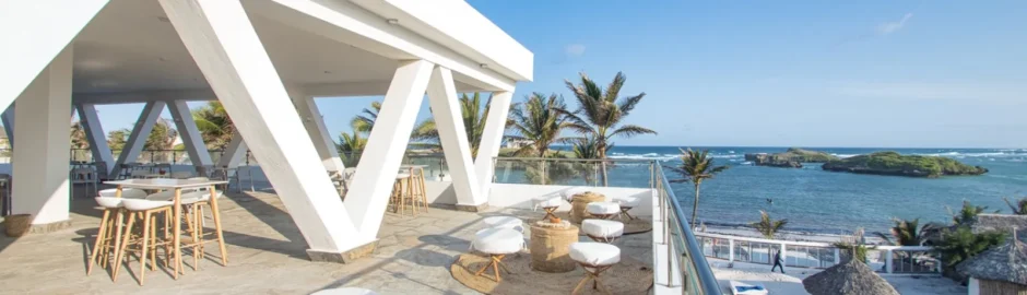 Terrazza coperta rialzata del Barracuda Inn Resort con pavimento in legno e una ringhiera protettiva, che offre una spettacolare vista panoramica sul giardino tropicale di palme, sulla spiaggia bianca sottostante e sulle acque azzurre di Watamu.