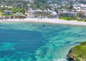 Fotografia aerea scattata con drone che mostra l'intero complesso del Barracuda Inn Resort a Watamu, evidenziando la piscina centrale azzurra, i curati giardini tropicali e le strutture bianche con i tetti in makuti affacciate direttamente sull'oceano.