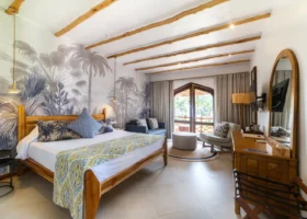 Interno di una luminosa camera da letto del resort Bluebay Beach con letto in legno, travi a vista sul soffitto e parete decorata con motivi tropicali