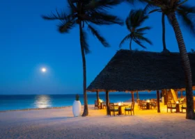 Tavoli illuminati sotto una grande struttura in makuti sulla spiaggia di Zanzibar di notte con la luna piena che si riflette sul mare