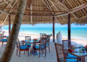 Tavoli in legno apparecchiati all'ombra di un patio in paglia sulla sabbia bianca con vista diretta sul mare cristallino di Zanzibar