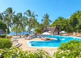Piscina dal profilo sinuoso circondata da lastre di pietra naturale e lettini in legno, incorniciata da alte palme da cocco affacciate sull'Oceano Indiano azzurro; capanne dal tetto in makuti e cespugli di fiori bianchi delimitano la scena sotto un cielo limpido.