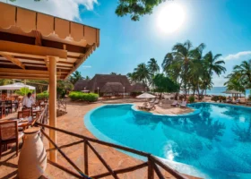 Terrazza ristorante con tavoli in legno apparecchiati e tettoia ombreggiante, affacciata su una grande piscina dalle acque turchesi; un cameriere in divisa bianca prepara i tavoli mentre palme da cocco e capanne dal tetto in makuti conducono lo sguardo verso il mare all'orizzonte.