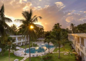 Tramonto dorato sul resort: il sole filtra tra le fronde di una palma da cocco proiettando un riflesso a stella, mentre la piscina dalla forma libera specchia il cielo arancione tra prati curati, vialetti a pietre tonde ed edifici bianchi in stile arabeggiante.