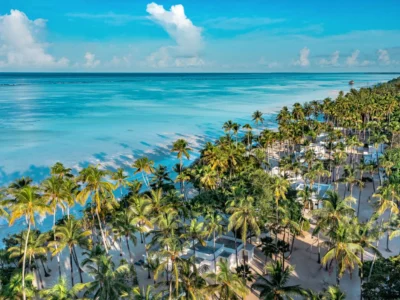 Veduta aerea panoramica del resort Diamonds Mapenzi Beach a Zanzibar, che mostra i bungalow bianchi immersi in un fitto palmeto tropicale lungo una spiaggia di sabbia chiara e le ampie sfumature turchesi dell'oceano Indiano sotto un cielo azzurro.