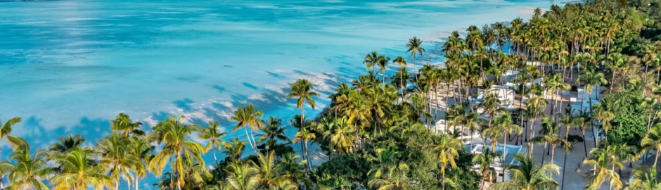 Veduta aerea panoramica del resort Diamonds Mapenzi Beach a Zanzibar, che mostra i bungalow bianchi immersi in un fitto palmeto tropicale lungo una spiaggia di sabbia chiara e le ampie sfumature turchesi dell'oceano Indiano sotto un cielo azzurro.