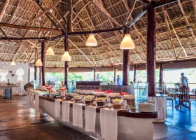 Area buffet del ristorante principale del Diamonds Mapenzi Beach a Zanzibar, allestita all'interno di una grande struttura con alto soffitto in legno e paglia makuti, caratterizzata da un lungo bancone a forma di barca ricoperto di piatti pronti.