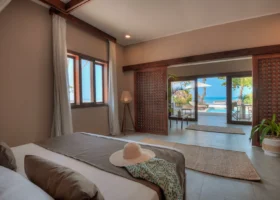 Interno di una lussuosa camera da letto del Diamonds Mapenzi Beach con letto matrimoniale in primo piano ornato da un cappello di paglia, pavimento in piastrelle scure e grandi porte scorrevoli in legno che aprono sul patio privato con vista mare.