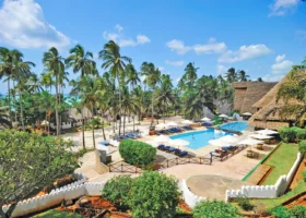 Ampia piscina all'aperto circondata da lettini prendisole e ombrelloni bianchi nel giardino tropicale del Diamonds Mapenzi Beach a Zanzibar, con alte palme da cocco sullo sfondo e le strutture principali del resort con tetti in makuti.