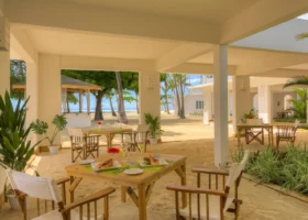 Terrazza coperta del ristorante del Diamonds Mapenzi Beach con pavimento in sabbia fine e tavoli in legno pronti per il pranzo, decorati con fiori rossi e calici, affacciati direttamente sulla spiaggia e sul giardino di palme esterno.