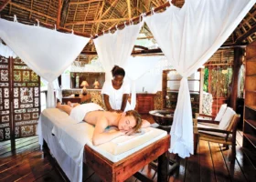 Interno della spa del Diamonds Mapenzi Beach a Zanzibar, dove una terapista in divisa bianca esegue un massaggio sulla schiena di una cliente distesa sul lettino, protetta da tende e zanzariere bianche sotto una struttura in legno rustico.