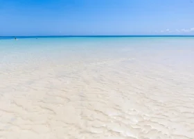 Dettaglio ravvicinato delle increspature della sabbia bianca finissima sommerse da pochi centimetri di acqua marina limpidissima e trasparente al Garoda Resort. L'orizzonte piatto mostra le diverse gradazioni di azzurro e turchese dell'oceano che si fondono con un cielo tropicale completamente sereno e privo di nuvole.