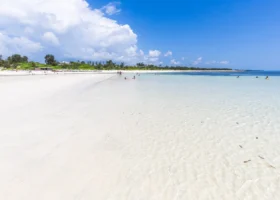 Inquadratura ad ampio raggio della celebre lingua di sabbia bianca che si estende nell'oceano Indiano di fronte al Garoda Resort a Watamu. L'acqua marina è trasparente e cristallina a riva, sfumando verso il blu cobalto in lontananza. Sullo sfondo si nota la costa tropicale con vegetazione lussureggiante e bagnanti che si rilassano sotto un cielo parzialmente nuvoloso.