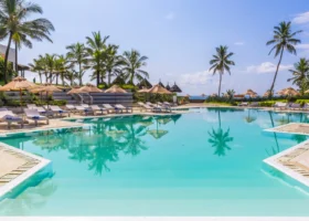 Grande piscina d'acqua dolce del Garoda Resort a Watamu, circondata da un'area solarium attrezzata con lettini prendisole bianchi e ombrelloni in paglia. Le alte palme da cocco si riflettono sulla superficie specchiata e turchese dell'acqua, delineando un contesto esotico sotto un cielo azzurro e luminoso.