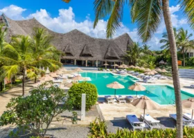 Veduta dall'alto dell'area centrale del Garoda Resort a Watamu, che mostra la grande piscina sagomata circondata da ombrelloni e lettini. Sullo sfondo si impone la struttura principale del resort, caratterizzata da grandi tetti tradizionali africani in makuti intrecciato, immersi in un giardino tropicale curato ricco di palme e piante ornamentali.