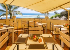 Terrazza panoramica coperta del ristorante all'aperto del Garoda Resort a Watamu. I tavoli in legno chiaro sono apparecchiati con runner scuri, posate e centrotavola di fiori tropicali colorati. La balaustra curva in muratura si affaccia direttamente sulla spiaggia e sulle barriere coralline visibili grazie alla bassa marea.