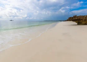 Tratto di spiaggia bianca e deserta adiacente al Garoda Resort a Watamu, caratterizzato da una imponente formazione rocciosa e scogliera corallina fossile sulla destra. Piccole onde lambiscono la battigia di sabbia soffice, mentre all'orizzonte un isolotto sperduto si staglia sotto un cielo carico di nuvole e schiarite.
