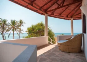 Patio coperto e terrazza panoramica dell'ingresso di una camera del Garoda Resort a Watamu. La struttura presenta un soffitto con travi in legno scuro a vista, colonne portanti bianche e un pavimento in pietra chiara. Sul balcone è posizionato un grande divano circolare in vimini intrecciato con cuscini bianchi, che si affaccia sul mare cristallino e sulle palme da cocco.