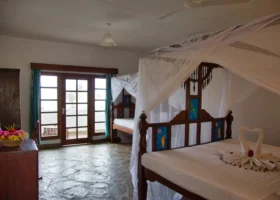 Interno di una camera da letto del Jacaranda Beach Resort a Watamu, arredata in stile locale con pavimentazione in pietra lucida, grande letto matrimoniale in legno scuro dotato di zanzariera a baldacchino bianca e asciugamani piegati a forma di cigno, affiancato da un secondo letto singolo e un'ampia porta finestra in legno che conduce all'esterno.