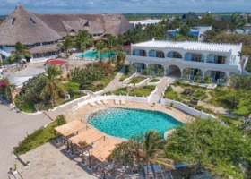 Veduta panoramica dall'alto del complesso centrale del Jacaranda Beach Resort in Kenya, che mostra una grande piscina azzurra di forma sinuosa circondata da vialetti in pietra, prati verdi curati, bouganville rosa fiorite e strutture alberghiere bianche in stile arabo-moresco con caratteristici tetti in makuti.