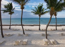 Area relax sulla spiaggia privata del Jacaranda Beach Resort a Watamu, caratterizzata da file ordinate di lettini prendisole in legno bianco posizionati sulla sabbia chiara di fronte al mare calmo, incorniciati da alte palme tropicali che si stagliano contro un cielo parzialmente nuvoloso.