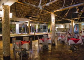 Interno dell'ampia sala ristorante del Jacaranda Beach Resort a Watamu, illuminata di sera da luci calde e caratterizzata da un imponente soffitto con travi a vista e tetto in paglia makuti, colonne portanti rivestite in pietra spugna locale, pavimentazione lucida e tavoli allestiti con tovaglie rosse e bianche.