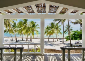 Veduta panoramica sull'oceano Indiano scattata dall'interno di una terrazza coperta del Jacaranda Beach Resort, incorniciata da una struttura geometrica bianca con travi in legno sul soffitto e parapetti a colonnine, che si affaccia direttamente su un giardino di palme e sulla spiaggia bianca sottostante.