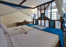 Interno di una camera da letto matrimoniale del Jumbo Resort a Watamu, caratterizzata da un grande letto in legno scuro provvisto di zanzariera a baldacchino bianca e azzurra, decorato con cigni fatti di asciugamani e fiori freschi, completato da una scrivania sullo sfondo.