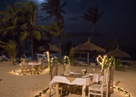 Allestimento serale sulla sabbia del Jumbo Resort a Watamu per una cena intima sotto la luna, con tavoli apparecchiati circondati da fili di luci calde a forma di cuore, ombrelloni in paglia makuti e alte palme mosse dal vento sullo sfondo.