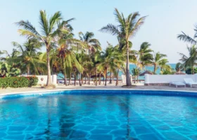 La grande piscina d'acqua dolce del Jumbo Resort a Watamu con pavimentazione in mosaico azzurro, circondata da un muretto bianco, lettini prendisole e slanciate palme tropicali da cocco che si stagliano contro un limpido cielo diurno.