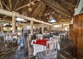 Interno del ristorante principale del Jumbo Resort in Kenya, caratterizzato da un maestoso soffitto alto con travi in legno e copertura in makuti, pavimentazione in pietra scura lucida e tavoli in legno massiccio apparecchiati con tovaglie rosse e bianche.