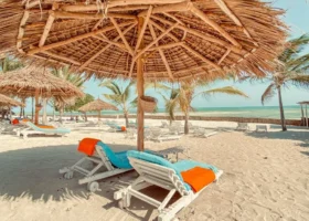 Area relax sulla spiaggia di sabbia finissima del Jumbo Resort a Watamu, attrezzata con lettini prendisole in legno dotati di teli mare azzurri e arancioni posizionati sotto un grande ombrellone in paglia intrecciata, con l'oceano sullo sfondo.