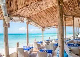 Tavoli apparecchiati con tovaglie blu e bianche all'interno di una terrazza panoramica coperta da una struttura in legno e makuti di fronte all'oceano
