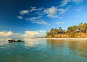 Una tipica imbarcazione in legno locale fluttua sulle acque turchesi e cristalline dell'Oceano Indiano a Diani Beach, Kenya. Sullo sfondo la spiaggia bianca del Kole Kole Beach Resort, costeggiata da una scogliera bassa coperta di palme e vegetazione rigogliosa sotto un cielo blu intenso con grandi nuvole bianche.