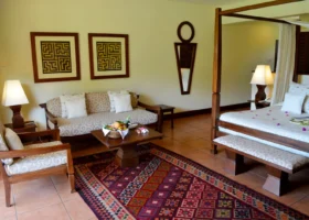 Interno spazioso di una camera deluxe del Kole Kole Beach Resort arredata in stile tradizionale africano. Sulla destra è posizionato un letto matrimoniale a baldacchino in legno scuro con zanzariera, mentre sulla sinistra si trova un'area relax con divano e poltrona coordinati, tavolino in legno con cesto di frutta fresca, quadri etnici alle pareti e pavimento in piastrelle di cotto.