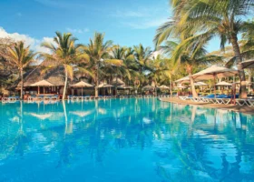 Ampia piscina esterna circolare adagiata in un lussureggiante giardino tropicale costellato di palme da cocco. Il perimetro della piscina è attrezzato con numerosi lettini prendisole dotati di cuscini blu e grandi ombrelloni chiari. Sullo sfondo si scorgono i tetti in paglia makuti delle aree comuni del Kole Kole Beach Resort.