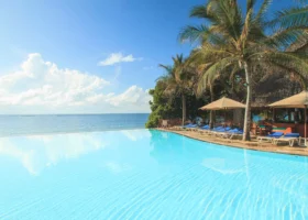 Grande piscina a sfioro con acqua azzurra e limpida che si fonde visivamente con l'orizzonte dell'Oceano Indiano. Sulla destra si sviluppa il solarium del Kole Kole Beach Resort pavimentato in pietra, attrezzato con lettini prendisole, teli mare blu, ombrelloni in tela beige, palme e la struttura del bar con tetto conico in makuti.