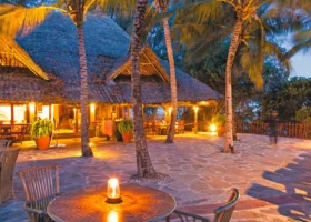 Terrazza esterna del ristorante del Kole Kole Beach Resort pavimentata in pietra irregolare, allestita con tavoli e sedie in legno sotto le palme illuminate alla sera. Sullo sfondo si erge la struttura principale del ristorante con grande tetto in paglia makuti sfiorata dalle calde luci dorate dei lampadari interni.