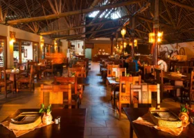 Interno del ristorante del Kole Kole Beach Resort illuminato da luci calde serali. La sala si caratterizza per un'imponente struttura a vista con travi in legno grezzo e alto soffitto in makuti. Tavoli e sedie in legno scuro sono disposti ordinatamente sul pavimento piastrellato, con lampade calde sospese che creano un'atmosfera accogliente.