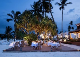 Tavoli apparecchiati su una pedana in legno illuminata da catene di luci tra le palme sulla sabbia bianca del Marijani Beach Resort al crepuscolo