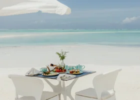 Un tavolo per due persone allestito sulla sabbia bianca sotto un ombrellone con pancake, frutta e tazze di caffè di fronte al mare del Marijani Beach Resort
